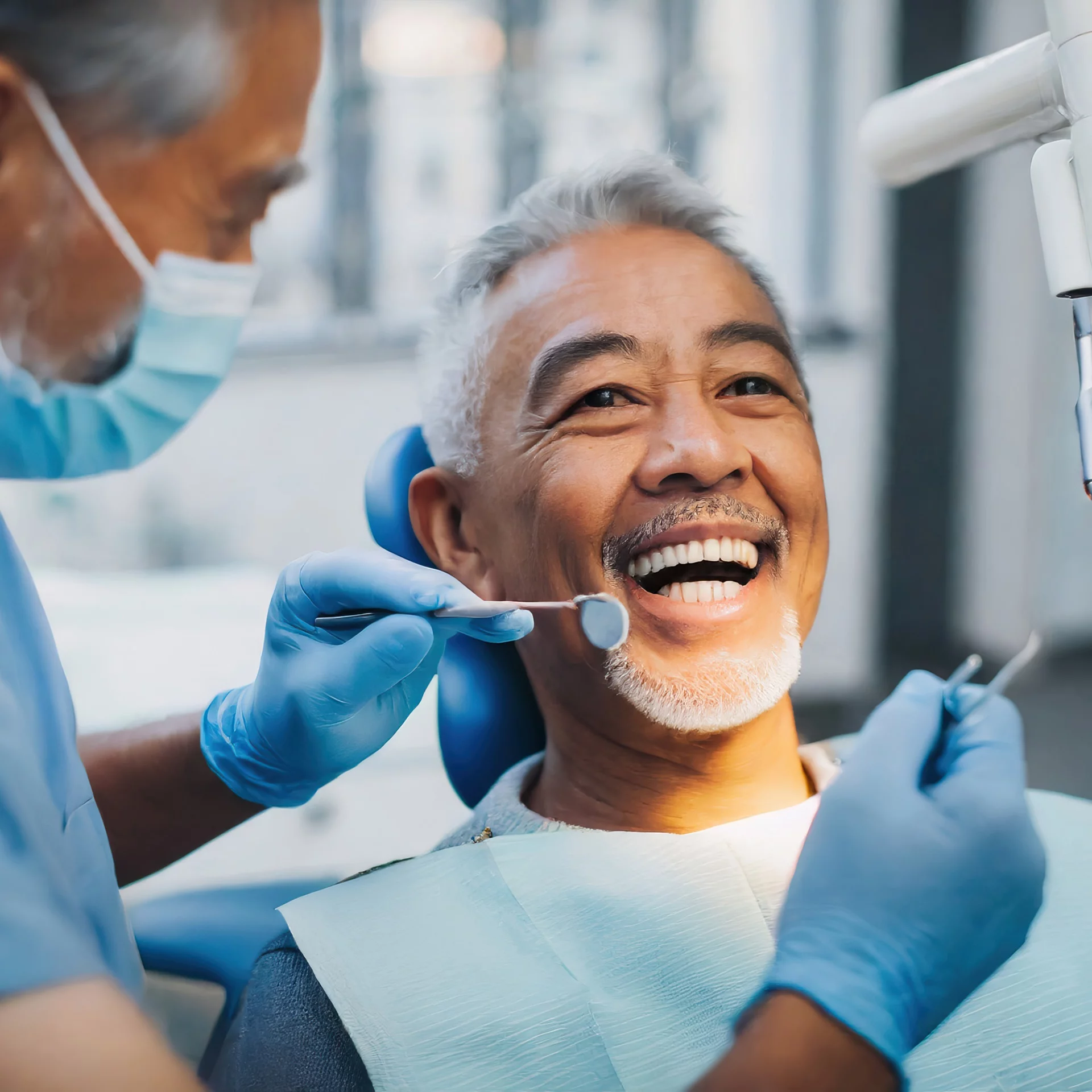 An elderly man with grey hair and a beaming smile is being attended to by a dentist wearing a mask and blue gloves. The dentist is examining the man's teeth with a dental mirror, indicating a routine dental check-up. The patient appears to be in a good mood and comfortable with the dental procedure.