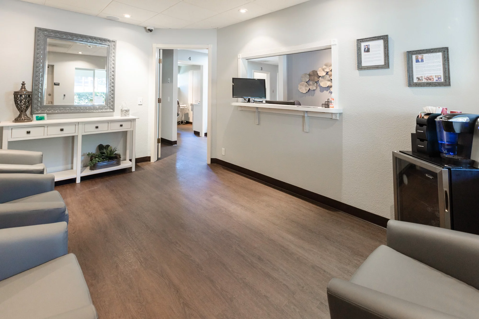The lobby of Placerville Dental Group is welcoming and elegantly appointed, featuring a contemporary design with comfortable gray chairs, wood-style flooring, and a well-organized reception area. The wall is adorned with informational frames, and the space is completed with a large ornamental mirror and a vase, creating a serene waiting experience. Open doors invite patients further into the practice, indicating a transition from the calming lobby to the professional care areas.