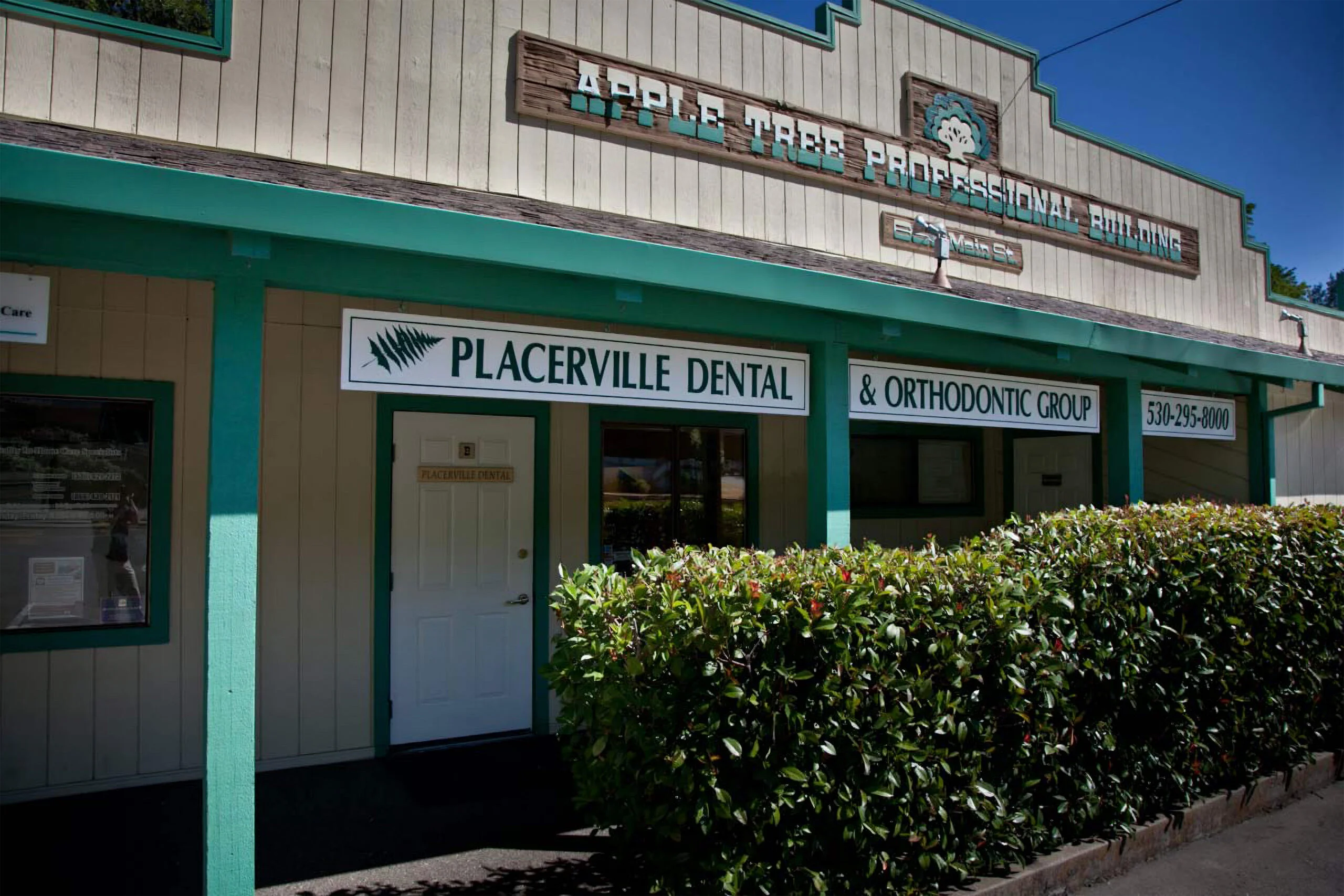 Exterior view of Apple Tree Professional Building, with signs for Placerville Dental & Orthodontic Group. The building features a beige facade with teal accents and a wooden signboard. Lush greenery in the form of trimmed hedges is visible in the front. Contact number 530-295-8000 displayed.