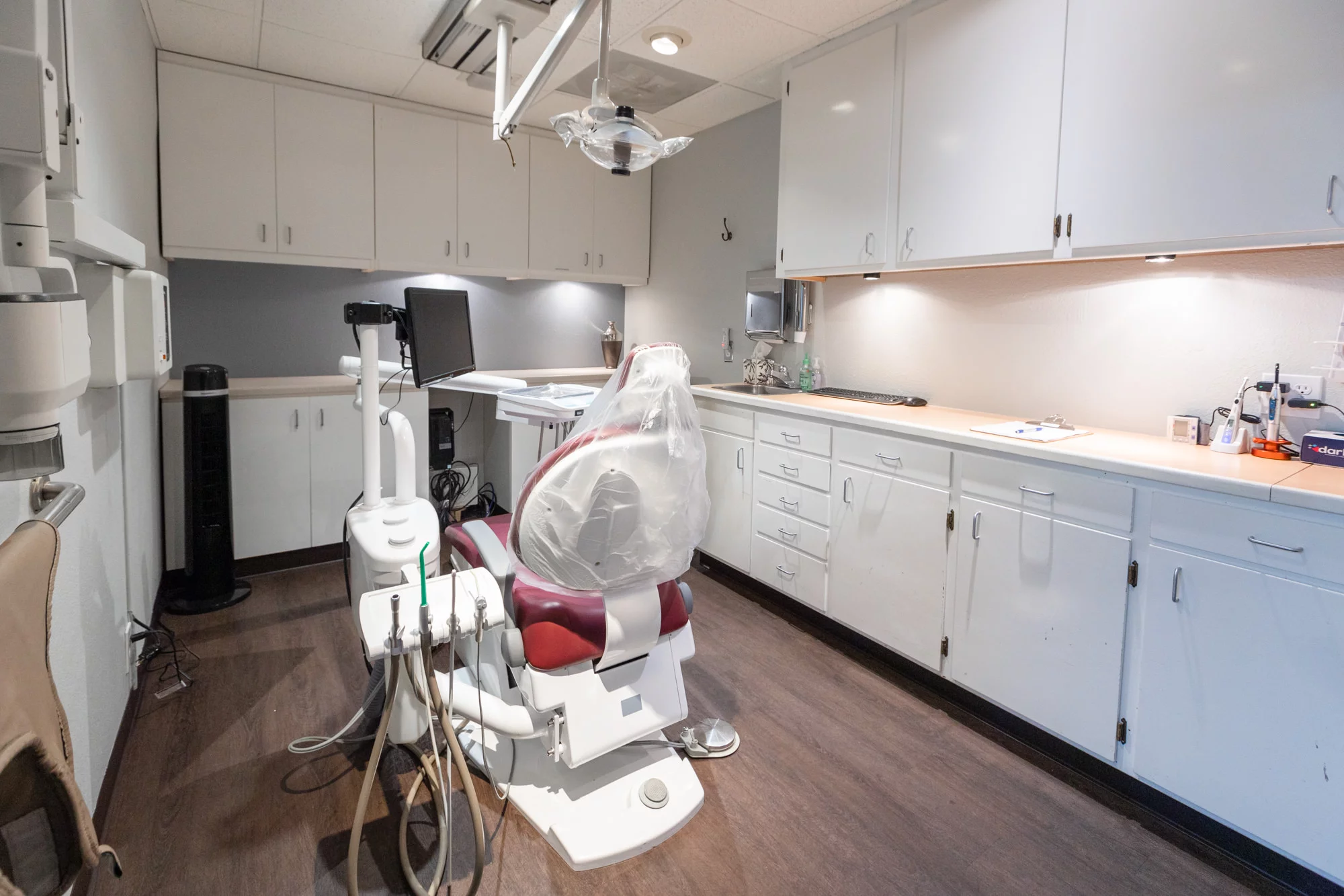 A fully equipped dental operatory with a maroon and white dental chair at the center, covered with a plastic sheet for hygiene. The room has upper cabinetry for storage, under-cabinet lighting illuminating a countertop workspace, and a sink area. Dental equipment and a computer workstation are present, with a dental X-ray machine on the left. The floor is finished with dark wood laminate, contributing to the professional and clean appearance of the space.