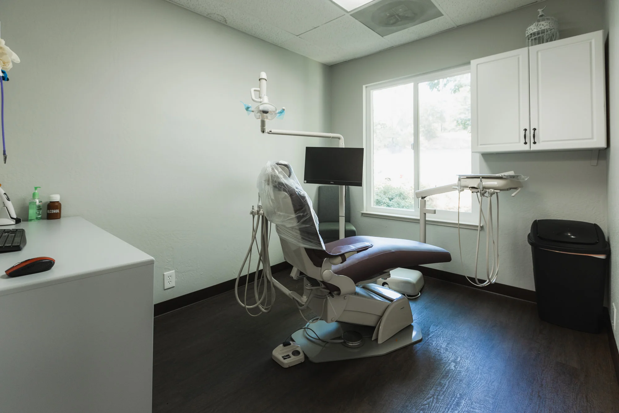 A clean and organized dental examination room featuring a purple dental chair with a plastic cover, positioned next to a large window for ample natural lighting. The room includes a white countertop with dental supplies, an overhead dental light, and a wall-mounted cabinet for additional storage. A computer monitor faces the chair for displaying patient information or X-rays.