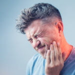 A middle-aged man with graying hair, squinting and grimacing in pain, holds his cheek and jawline suggesting a toothache or jaw discomfort. He wears a casual blue t-shirt, set against a soft blue gradient background.