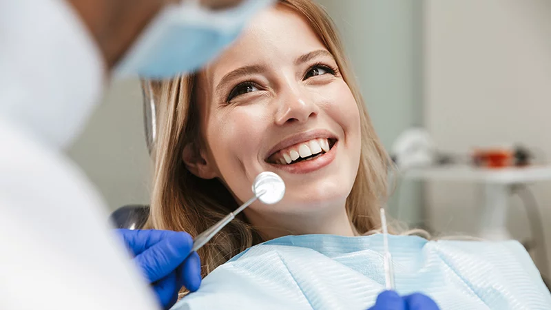 A cheerful woman with blonde hair, lying in a dental chair, smiles up at her dentist, who is out of frame. The dentist's hands, gloved in blue, are holding a dental mirror and a probe near the woman's mouth. The scene is set in a dental office, indicated by the chair and clinical surroundings.
