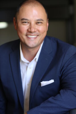 A professional headshot of Dr. Steele, featuring his pleasant smile and composed demeanor. He is dressed in a sharp navy blue suit complemented by a crisp white shirt, with a white pocket square adding a touch of elegance to his suit jacket. The photo is thoughtfully cropped to focus on his upper body and face, capturing every detail in high definition. The background is softly blurred, ensuring that the emphasis remains on his engaging expression and professional poise.