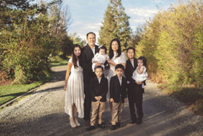 Dr. Brian Steele and Nancy, alongside their six children, in semi-formal wear, gathered on a gravel walkway amidst vibrant green foliage for a family photograph.