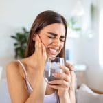 A young woman with long brown hair, wearing a white tank top holds a glass of ice water in her left hand while clenching her teeth and holding her face with her right hand. She appears to be in pain. Behind her sits a white couch and desk with a plant on it. The room is bright and light.