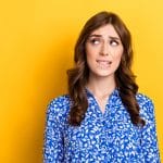 ALT text: A woman with wavy brown hair wearing a blue blouse with a white floral pattern looks off to the side with a nervous or uncertain expression, slightly biting her lip. The background is a solid bright yellow.