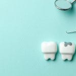 Dental concept image showing two metal dental tools—a mirror and a probe—lying on a light blue textured background beside three model teeth, with the middle tooth featuring a visible cavity or filling on the top surface.
