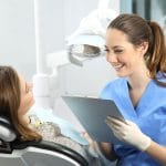 Smiling dental hygienist in blue scrubs holding a clipboard and talking to a female patient reclining in a dental chair, in a bright, modern dental office.