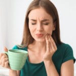 Woman with long brown hair wearing a green shirt holding a green ceramic mug and touching her cheek with a pained expression, indicating tooth or jaw discomfort.