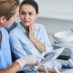 Dentist showing a dental X-ray to a concerned patient touching her jaw during a consultation.