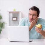 Man holding his cheek in discomfort while eating an apple at a desk with an open laptop.