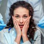 Anxious dental patient holding her face while gloved hands with dental tools approach during an exam.