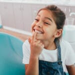 Young girl sitting in a dental chair, smiling and pointing to her front tooth in a bright clinic
