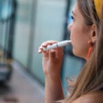 Person holding a slim white vape pen to their lips, seen in side profile indoors with soft background blur.