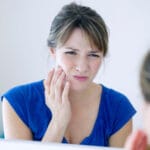 Woman touching her cheek and wincing in pain while looking in a mirror, suggesting toothache or jaw discomfort