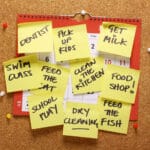 Corkboard calendar covered with yellow sticky notes listing tasks like dentist, pick up kids, get milk, and clean the kitchen