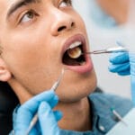 Dentist wearing gloves examines a patient’s open mouth with a mirror and dental scaler during an oral checkup