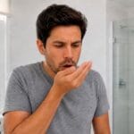 Man covering his mouth and checking breath in a bathroom mirror, concerned about possible bad breath