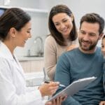 Dentist in a modern dental office reviews a clipboard with a smiling family of three, including a young girl seated beside her parents, during a friendly consultation.