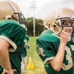 Young boys in green foothball uniforms putting on their helmets and mouthguards to prevent dental injury.